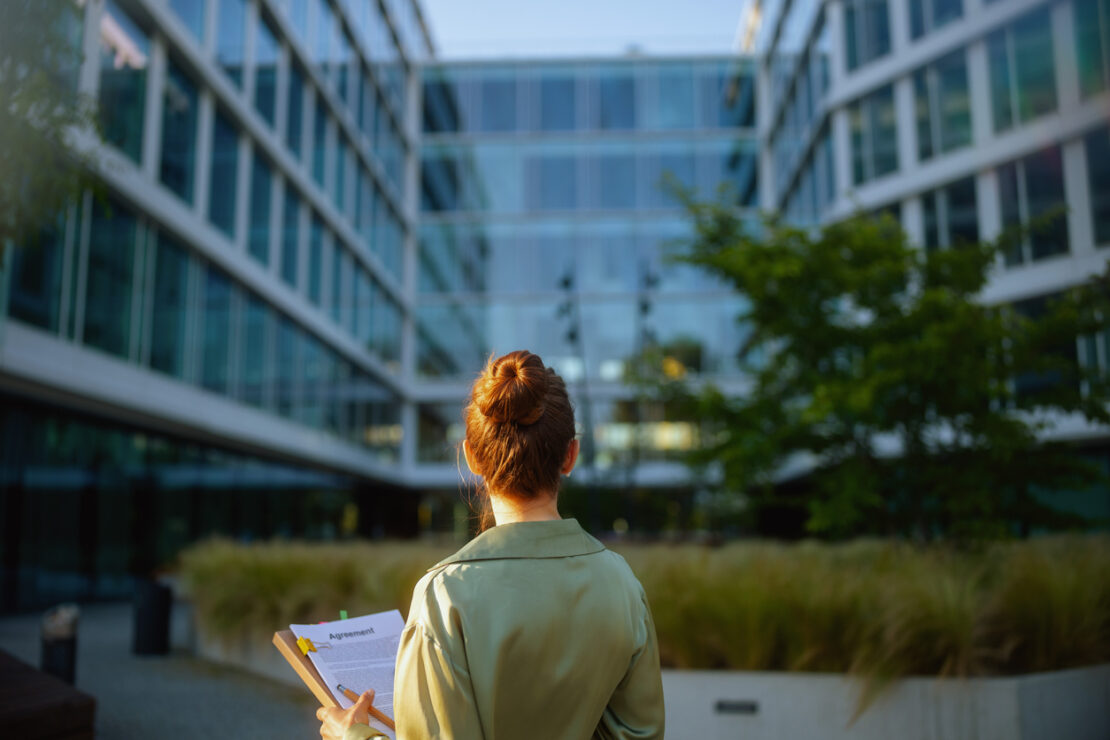 Seen from behind business woman near office building in green blouse with documents.